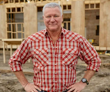 Man in a red plaid shirt standing at a construction site, wooden frame structure in the background.