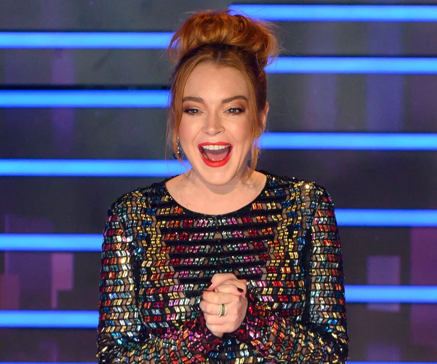 Lindsay Lohan on stage, in a colorful sequined dress, smiling and clapping hands, with a blue-lit background.