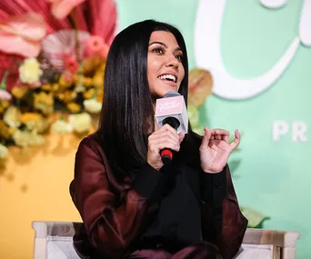 A woman speaking into a microphone at an event, with a colorful floral backdrop.