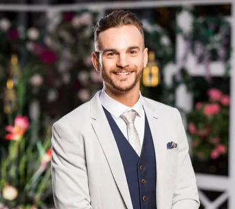 Man in a light suit with a vest and tie, smiling, standing in front of a floral background.