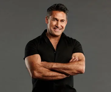 Man in a black shirt smiling with arms crossed against a gray background.
