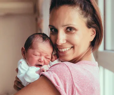 A mother lovingly holds her newborn baby, both smiling in a warm, well-lit room.