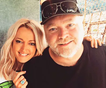 Two people smiling at the camera, one with long blonde hair and a ring, the other with a cap and sunglasses on head.