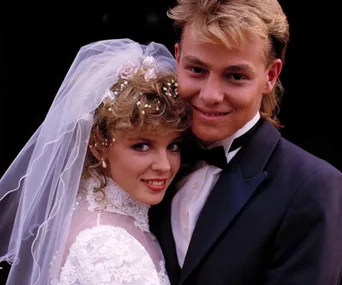A bride in a white lace gown and veil leans into a groom in a tuxedo, both smiling, in a classic wedding pose.
