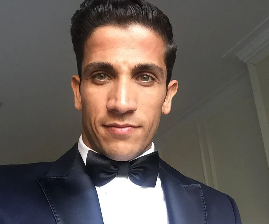 Man in a tuxedo and bow tie, close-up selfie, with neutral expression against a light background.