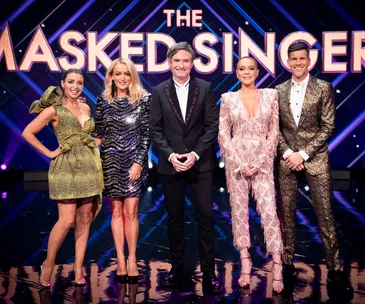 Judges of "The Masked Singer" stand on stage in glamorous outfits, with the show logo in the background.