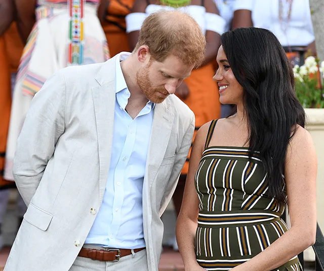 Two people are smiling at each other, one in a light suit and the other in a striped dress, outdoors.