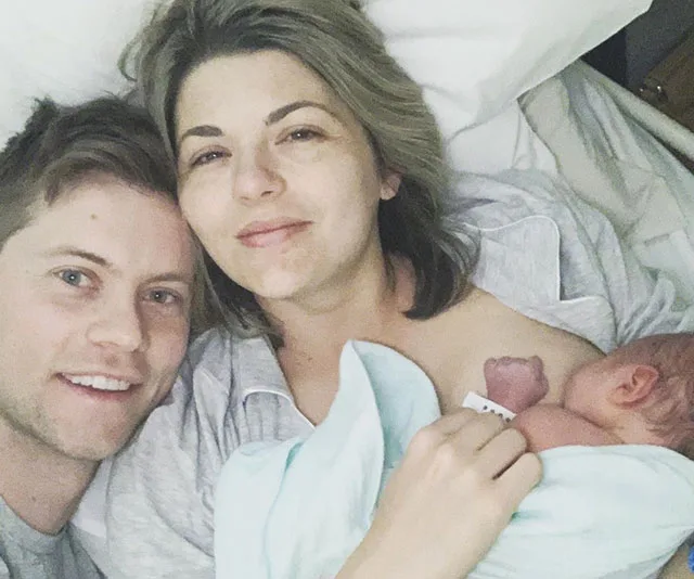 "Couple in hospital bed smiling with a newborn baby wrapped in a blue blanket."