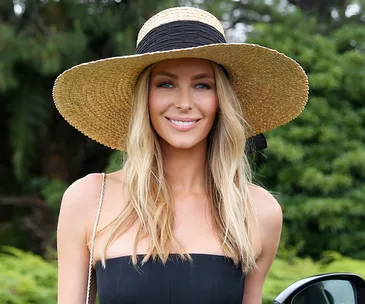 Woman in a straw hat with long blonde hair smiles outdoors, wearing a sleeveless top.