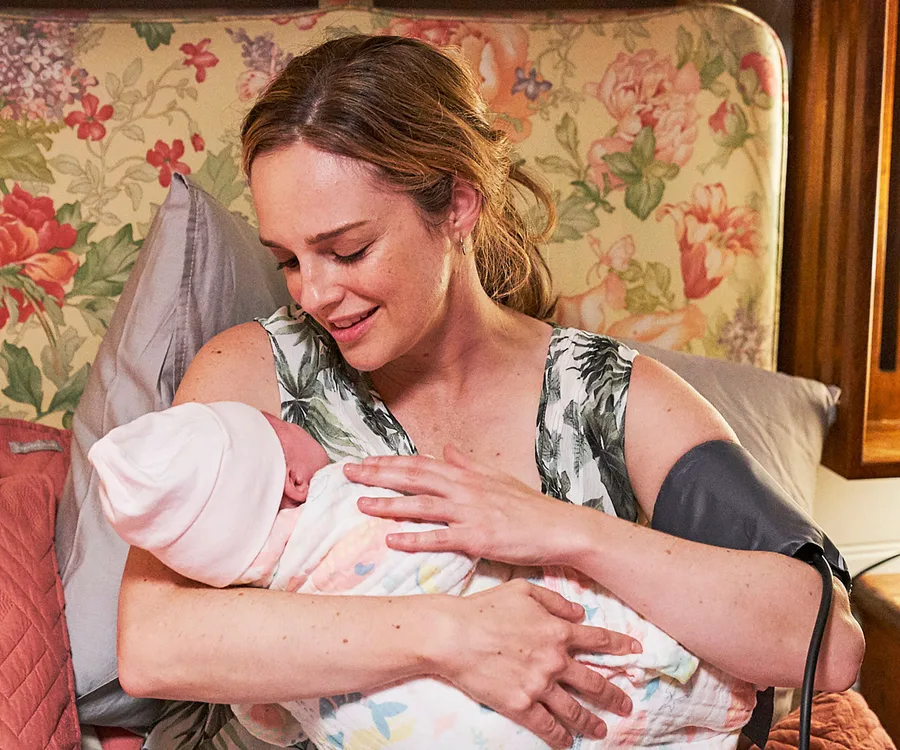 A woman with light brown hair smiles while cradling a newborn wrapped in a blanket, with a floral backdrop.