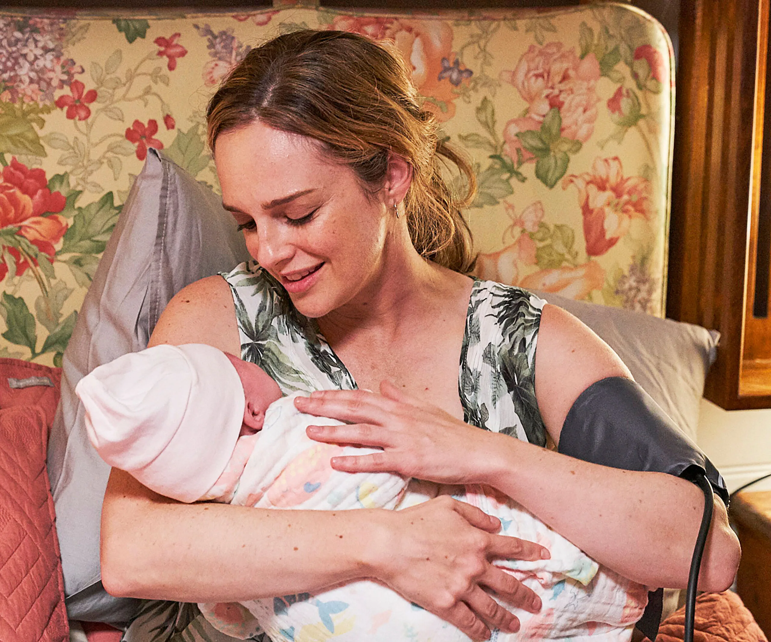 A woman with light brown hair smiles while cradling a newborn wrapped in a blanket, with a floral backdrop.