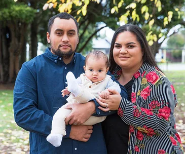 A family of three outdoors; a man and woman holding a baby, surrounded by trees and fallen leaves.