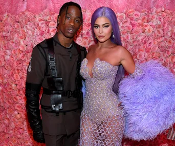 Two individuals posing in front of a pink floral backdrop, one in a dark outfit and the other in a sparkling dress with purple hair.