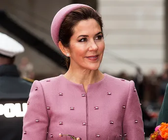 A woman in a pink outfit and hat smiles at an event, with a blurred background and people nearby.