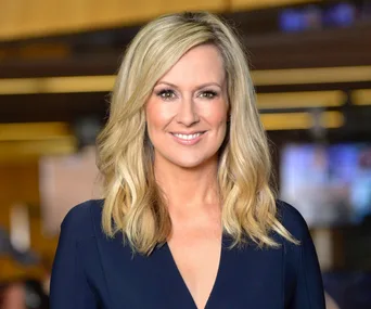 A woman with long blonde hair smiling, wearing a dark top, with a blurred background of an indoor setting.