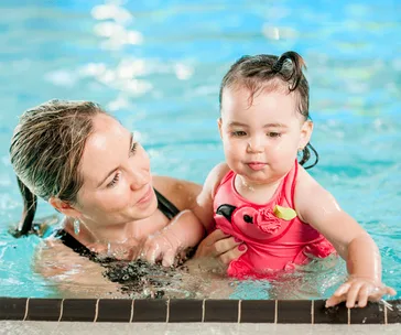 Mother and baby in a pool, baby in pink swimsuit, mother smiling and supporting her.