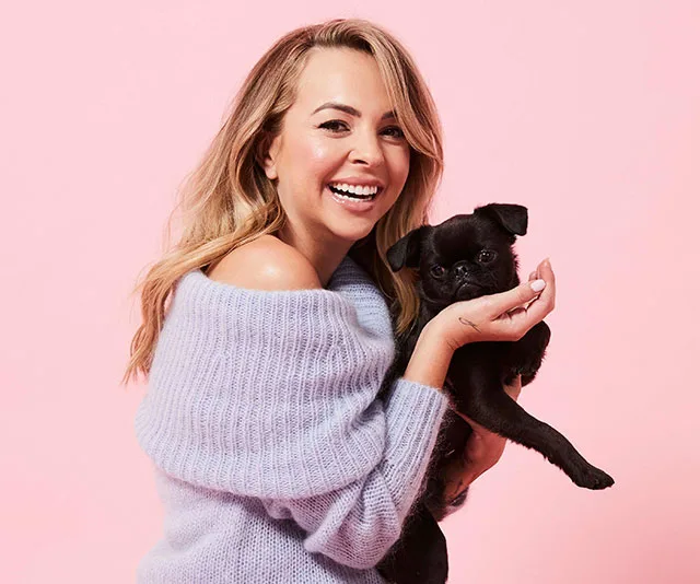 Woman in a purple sweater smiling and holding a small black dog against a pink background.
