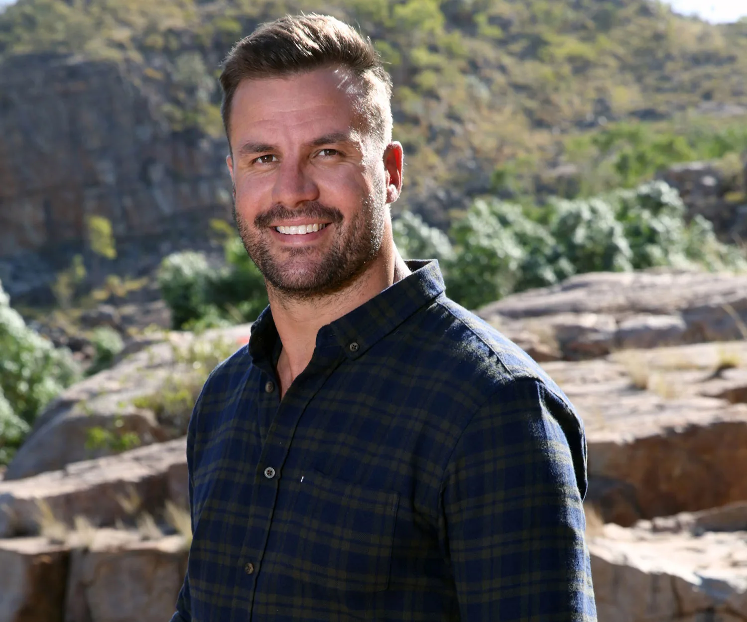 A smiling man in a plaid shirt stands outdoors with rocks and greenery in the background.