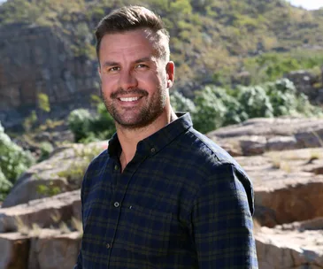A smiling man in a plaid shirt stands outdoors with rocks and greenery in the background.