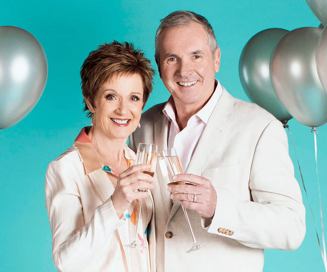 Two people, a woman and a man, smiling and holding champagne glasses, with silver balloons in the background.