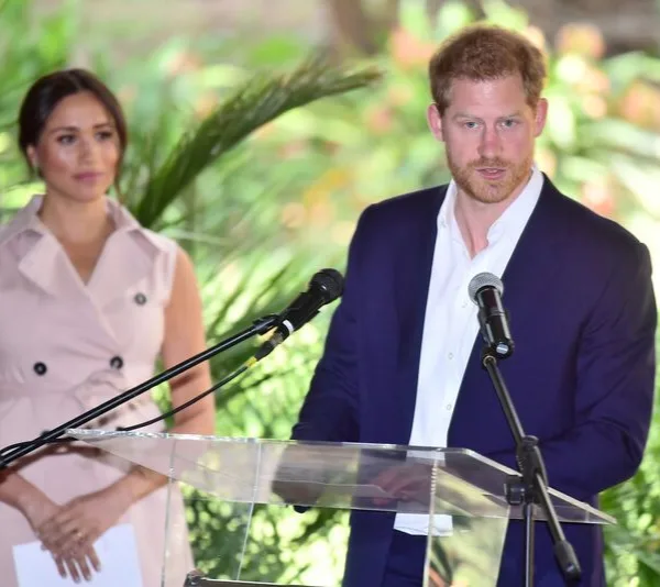Couple at an outdoor podium event. Individual in a blue suit speaks, while another in a pink dress stands nearby.