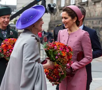 Princess in pink and an older woman in a purple hat smiling and exchanging bouquets outdoors.