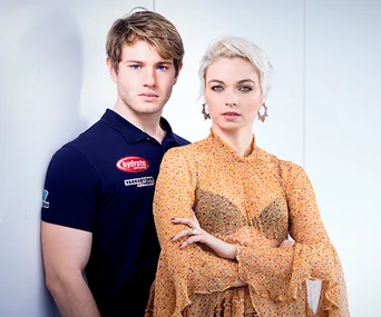 A man in a navy shirt stands beside a woman in a patterned blouse, both looking directly at the camera against a plain backdrop.
