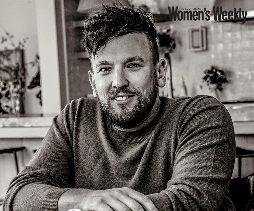 A man with short, dark hair and a beard, wearing a sweater, smiling while sitting in a cozy indoor setting.