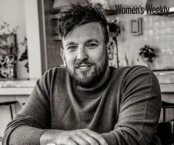 A man with short, dark hair and a beard, wearing a sweater, smiling while sitting in a cozy indoor setting.