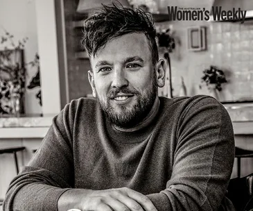 A man with short, dark hair and a beard, wearing a sweater, smiling while sitting in a cozy indoor setting.