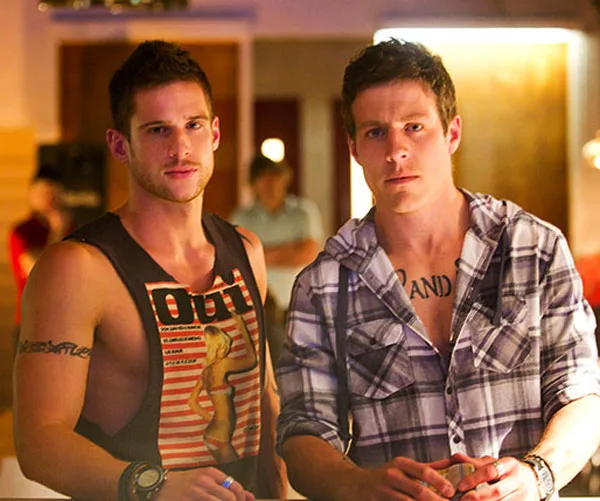 Two men with tattoos stand in a dimly lit room, both looking towards the camera; one in a tank top, the other in a plaid shirt.