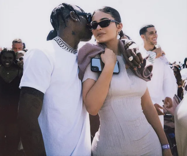 A couple embraces closely amidst a crowd, with both wearing sunglasses.