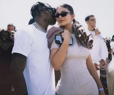 A couple embraces closely amidst a crowd, with both wearing sunglasses.