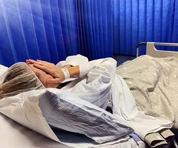 Person in hospital bed covering face with hands, wearing a hospital gown and cap, blue curtains in the background.