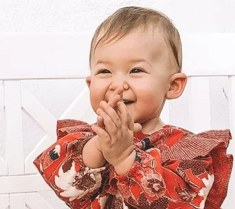 Baby in a red floral dress smiles and claps hands, sitting on a white chair.