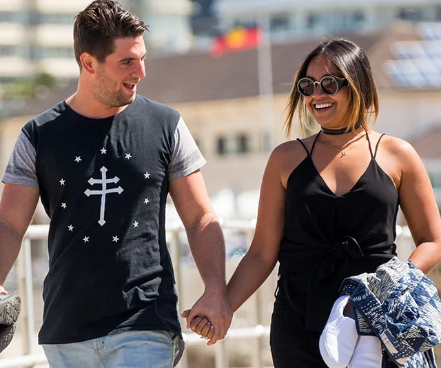 A couple holding hands and smiling at each other while walking outdoors on a sunny day.