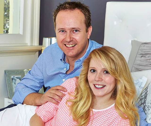A smiling couple sitting together on a bed in a bright, stylish room.