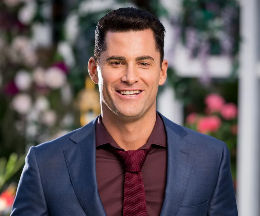 A smiling man in a navy suit and maroon tie, with a backdrop of blurred flowers and greenery.