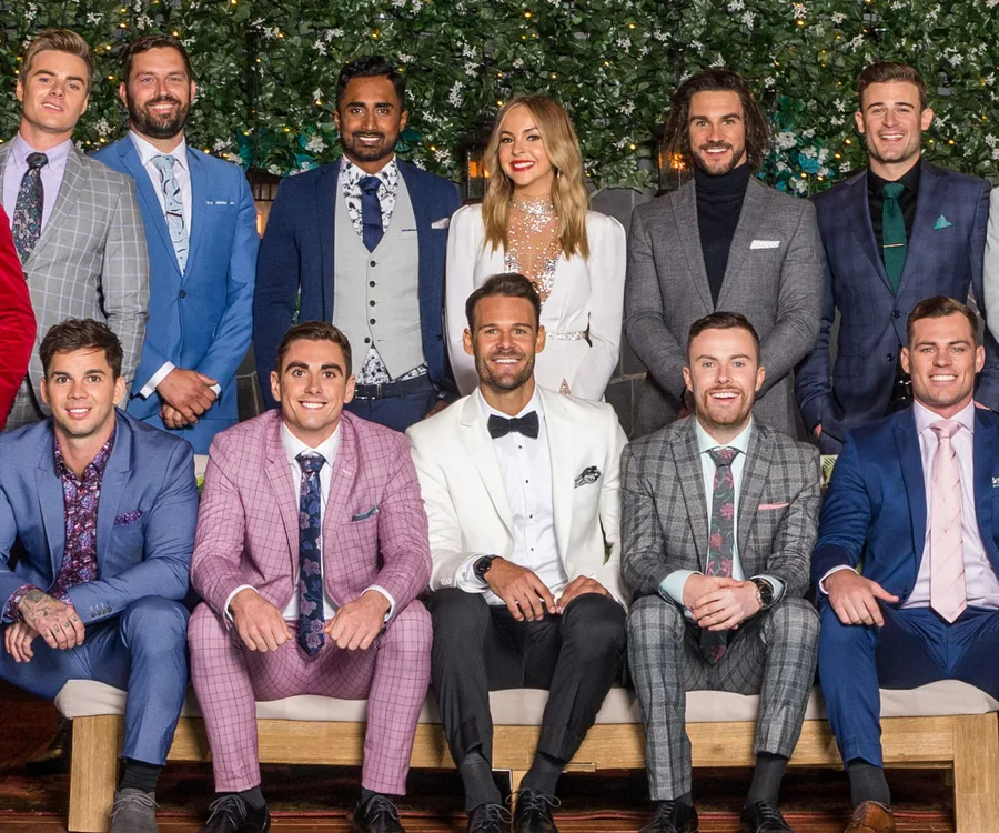 A group of men and one woman in formal attire pose for a photo, standing and sitting on a bench against a floral backdrop.