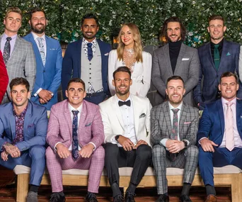 A group of men and one woman in formal attire pose for a photo, standing and sitting on a bench against a floral backdrop.