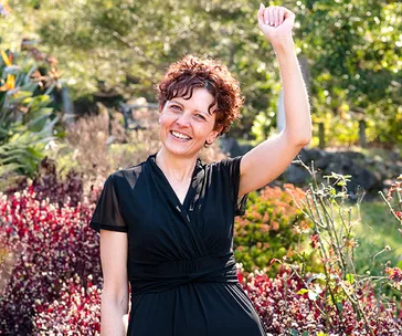 A woman in a black dress smiles and raises her arm in celebration, standing in a lush garden.