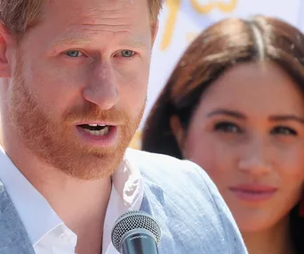 Prince Harry speaking into a microphone with Meghan Markle in the background.
