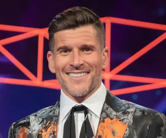 A smiling man in a colorful suit stands against a vibrant, geometric background.