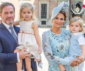 A royal family with a man, woman in blue dress and hat, and two young children in a courtyard setting.