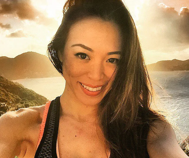 A woman smiles at the camera during a sunny day by the sea, surrounded by hills and sky.