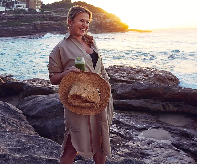 Woman in a beige dress holding a straw hat and green drink on rocky beach at sunset.