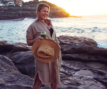 Woman in a beige dress holding a straw hat and green drink on rocky beach at sunset.