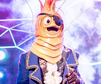 A costumed singer in a shrimp-like outfit with an eye patch, performing on stage with a vintage microphone.