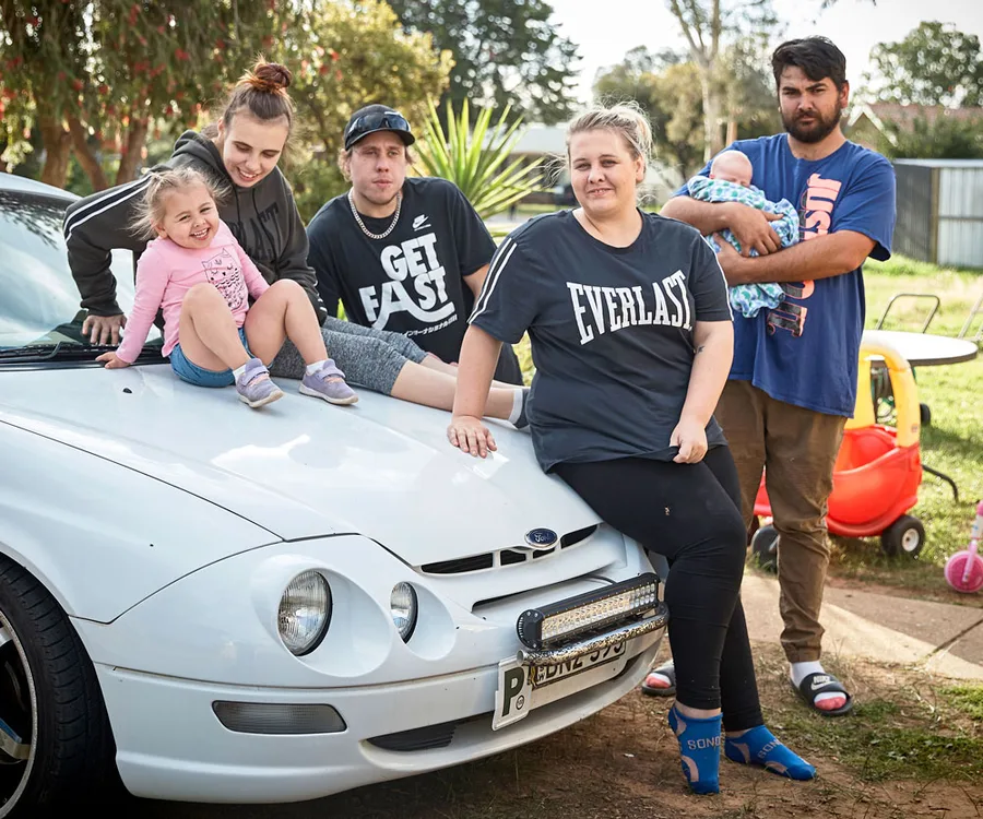 Group of five people, including two children, sitting and standing around a white car in a backyard setting.