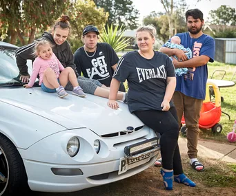 Group of five people, including two children, sitting and standing around a white car in a backyard setting.